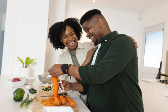 African American couple laughing, peeling carrots on kitchen island using peeler and cutting board