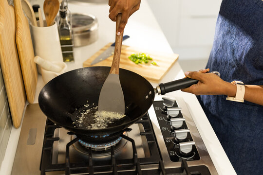 African woman wearing denim apron stirring garlic in black wok on gas stove in kitchen