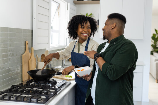 African American couple cooking in kitchen, woman in blue apron stirring wok while man holding bowl