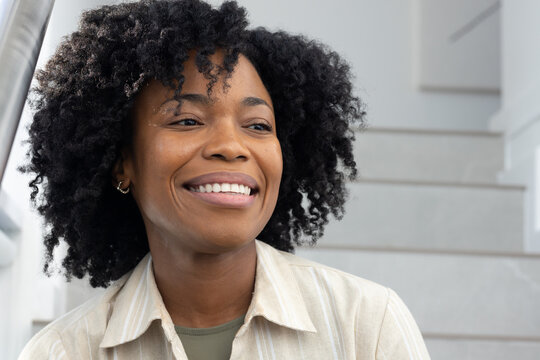 African woman smiling, looking right on stairs beside metal rail, wearing cream shirt, hoop earring