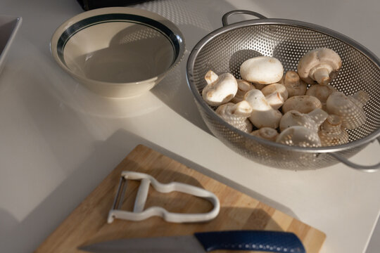 Metal colander is holding white button mushrooms on light counter with bowl and peeler in sunlight