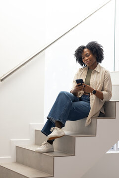 African American woman sitting on stairs near window, holding smartphone by handrail, copy space