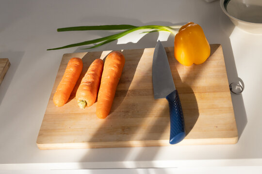 Wooden cutting board is sitting on white countertop near window, holding carrots, pepper