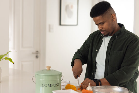 Adult African man preparing meal, slicing pepper and garlic on wooden board by compost bin