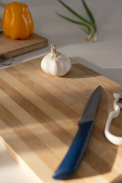 Whole garlic bulb sitting on diagonal bamboo board, blue-handled knife, yellow pepper and peeler