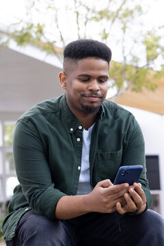 African American man sitting on bench in courtyard holding navy phone, wearing green shirt over tee