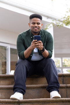 African American man sitting on stone porch steps holding blue smartphone wearing smartwatch shoes