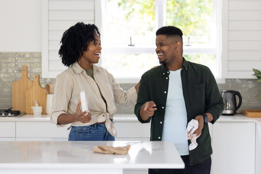 African American couple standing at white kitchen island holding bottle and dish towel, smiling
