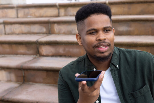African American man holding smartphone and speaking into device, sitting on stone steps