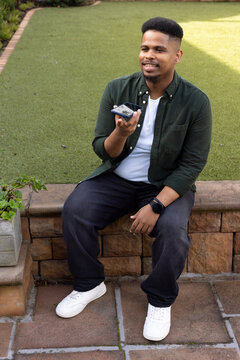 Adult African American man sitting on low stone wall at backyard patio, holding smartphone