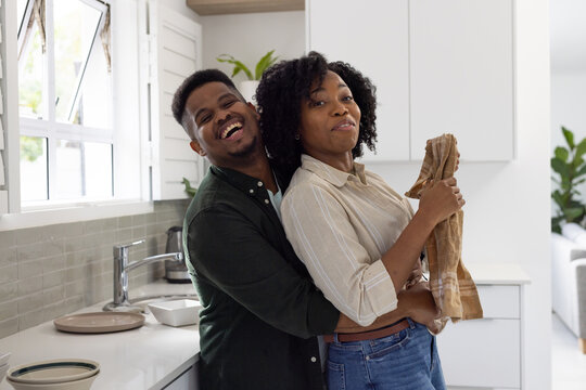 African couple hugging and laughing near kitchen sink with chrome faucet, woman holding brown towel