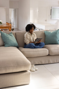 African American woman sitting on beige sofa in bright room wearing beige shirt using tablet