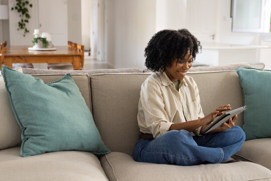 African woman sitting cross-legged on beige sofa with teal pillows, holding tablet at home in jeans