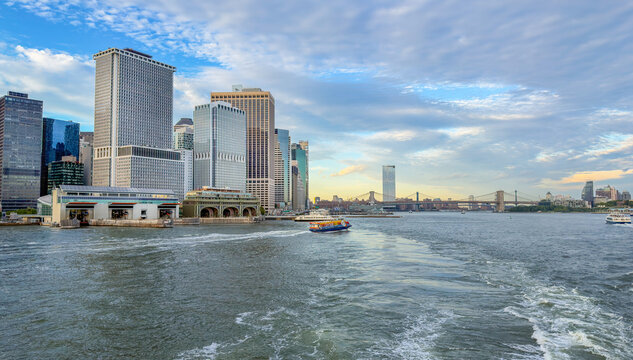 View of the Manhattan skyline from the East River with a ferry boat crossing the water under a cloudy blue sky in New York, New York, United States.