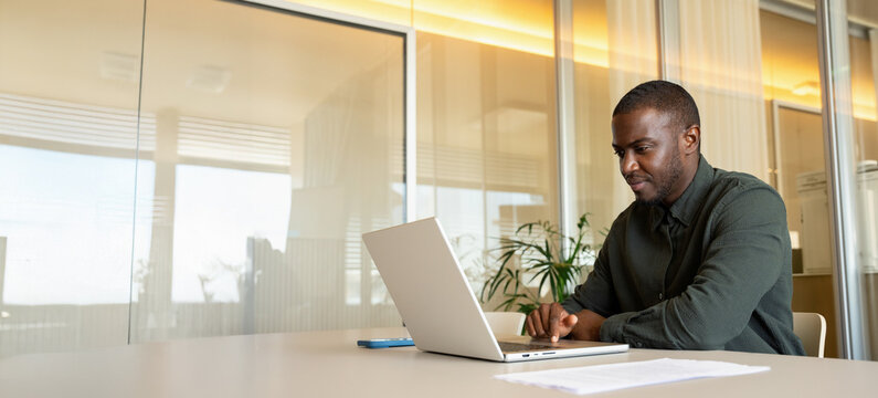 Web banner of african business man in 30s typing on laptop computer at office desk, black analyst reading online report inside coworking area, focused employee during working day. Copy space.