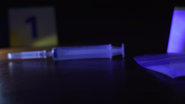 Panning from drug baggie at marker two to capped syringe at marker one under flashlight and flashing police lights, low angle close up