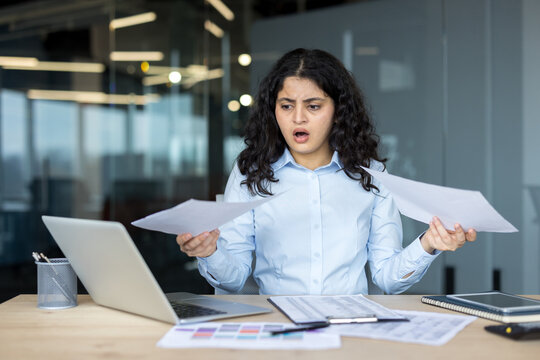 Stressed Businesswoman Working On Laptop And Analyzing Documents At Her Desk In A Corporate Office. The Professional Female Manager Is Feeling Overwhelmed While Reading Financial Analysis Reports.