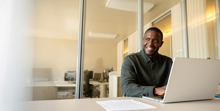 Web banner of smiling african business man in 30s sitting behind laptop at office table, friendly black specialist portrait looking at camera inside coworking area, corporate staff. Copy space.