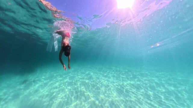 Stunning freediver in pink swimwear gliding through transparent turquoise ocean water toward sandy floor. Underwater tracking shot.