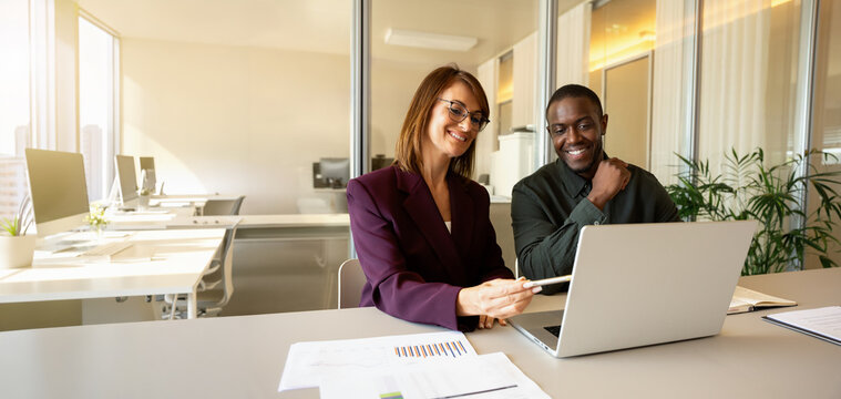 Web banner of business woman in 40s pointing pen at laptop screen beside smiling african man in 30s, diverse colleagues reading financial charts inside open office, corporate training. Copy space.