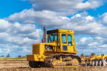 Agricultural view of yellow bulldozer cultivating soil with disc harrow under cloudy blue sky. © Todayphoto
