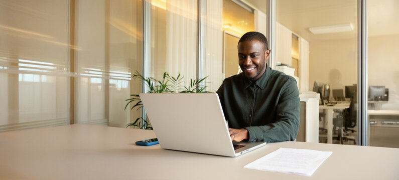 Web banner of african business man in 30s working on laptop computer at modern office desk, smiling black employee checking financial report, banker analysing market statistics online. Copy space.