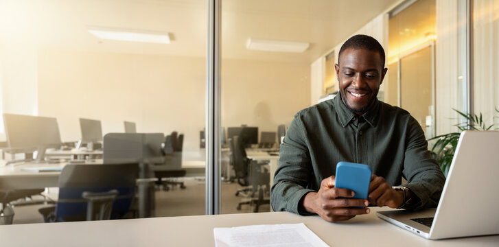 Web banner of african business man in 30s texting on smartphone phone at office desk beside open laptop, smiling black employee messaging client during working day, mobile banking. Copy space.