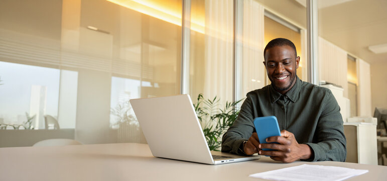 Web banner of smiling african business man in 30s scrolling on mobile phone beside laptop at office table, black professional checking online messages, fintech employee at daily work. Copy space.