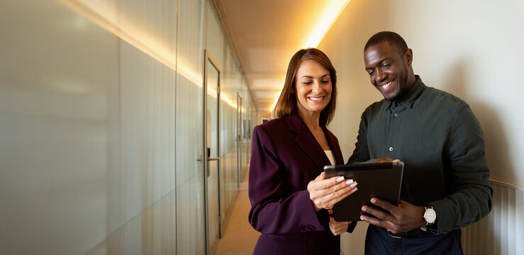 Web banner of multiracial business colleagues using digital tablet at office corridor, smiling woman in 40s and african man in 30s discussing online report, corporate teamwork for company. Copy space.