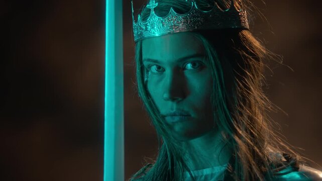 Portrait of young Caucasian woman wearing crown, holding glowing sword while looking at camera. Footage suits fantasy gaming marketing, cinematic trailers, and cosplay advertising
