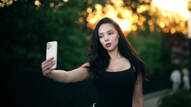 Beautiful attractive young woman taking selfie on smartphone in park at sunset. Pretty brunette female posing and holding mobile phone outdoors with bright lens flare background