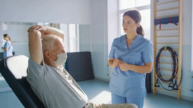 Guiding therapist in scrubs urging masked patient raising arms on recliner for motion in rehab