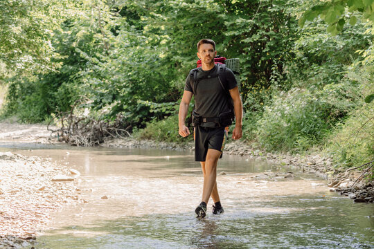 Handsome man hiker walking through a shallow forest stream with a backpack during a summer adventure