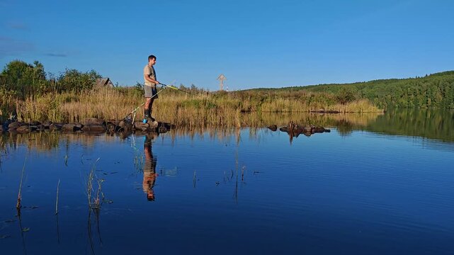 Man fishing while standing on a reedy riverbank with green hills reflecting in the calm water at golden hour, tranquil angling, outdoor leisure, riverside serenity.