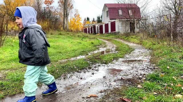 Cute little toddler child kid boy playing outside in boots in muddy puddle after rain in garden,home backyard at autumn. Carefree childhood. Moody fall weather forecast