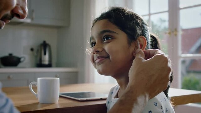 Reaching father tucking girl's hair, placing, securing ear device in kitchen, wearing floral top