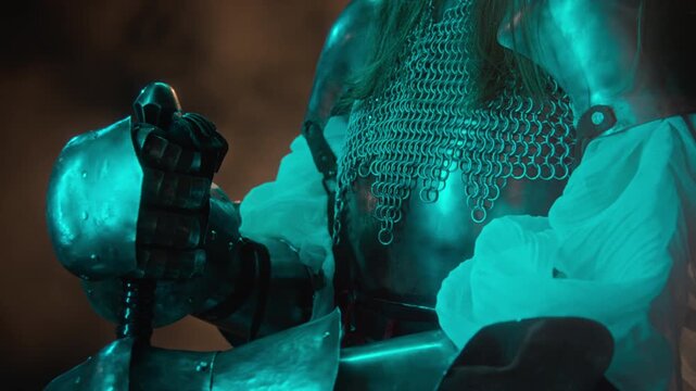 Young adult female knight wearing steel armor holding sword hilt, camera tilting up. Footage suitable for historical documentaries, fantasy gaming promos, or medieval trailers