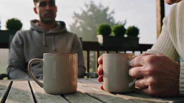 Senior man in knit sweater lifting mug and sipping during conversation on porch, with beige mugs