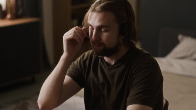 Caucasian young adult man putting on headphones and resting head on hand while listening to music indoors. Clip suits mental health campaigns, audio streaming ads, or remote work wellness content