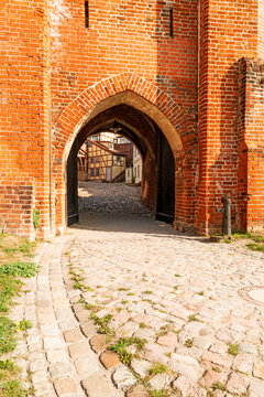 Historic brick gate with a pointed arch passage and view toward half timbered houses, photographed in vertical format.