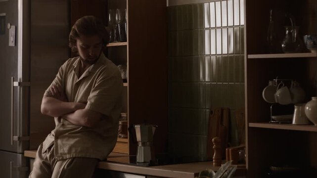 Caucasian young adult man waiting for coffee to brew in moka pot while leaning against kitchen counter. Footage suitable for daily wellness, morning routine projects and beverage advertisements