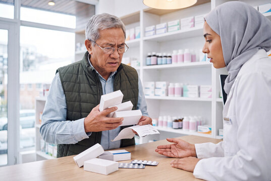 Confused senior East Asian man holding several medicine boxes and prescription, asking compassionate Middle Eastern pharmacist in hijab for help at pharmacy counter. Medication worries