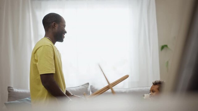 African American father engaging in playful wooden sword fight with cheerful child son at home. Scene fits fatherhood branding, parenting campaigns, and family lifestyle projects