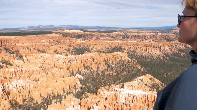 Tourist blonde woman looking at stunning panoramic view of bryce canyon amphitheater, filled with unique orange hoodoos and green pine trees under a clear sky