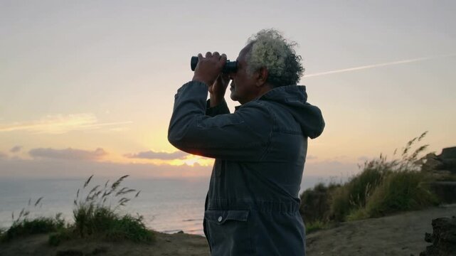 Lifting binoculars senior man in parka on grass cliff, adjusting grip scanning ocean as sun sinking
