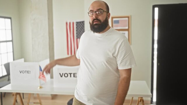 Man with beard and glasses points at a ballot box and shows open palms in a polling building with flag and vote booths visible; civic duty uncertainty.