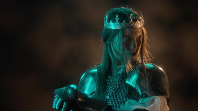 Caucasian young woman wearing medieval knight armor and royal crown turning head cautiously. Footage suitable for fantasy films, video game trailers, and historical reenactment projects