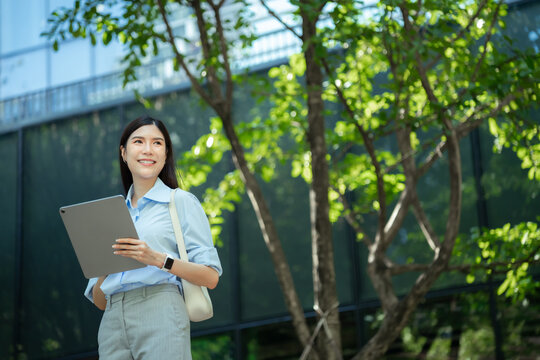 Wide shot of an asian businesswoman holding a tablet computer in a green city park, with sunlit leaves filling the right side of the frame.