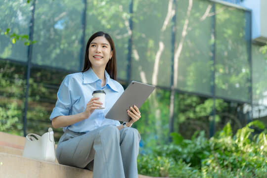 Asian businesswoman sits on a low wall outdoors with a tablet computer and a takeaway coffee cup, her handbag beside her during a work break.