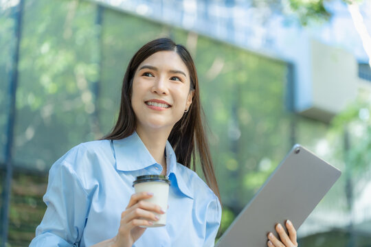 Close view of an asian businesswoman holding a paper coffee cup and a tablet computer outdoors, looking up with a relaxed smile.